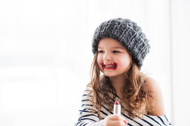 Ein kleines, fröhliches Mädchen im gestreiften T-Shirt, das zu Hause einen Hut trägt und Lippenstift aufträgt, kopiert Raum. - HPIF30637