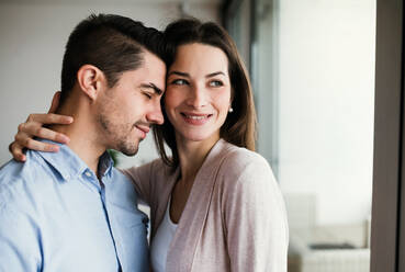 Young happy couple in love standing by the window at home, hugging. Copy space. - HPIF30689