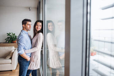 Young happy couple in love standing by the window at home, hugging. Copy space. - HPIF30691