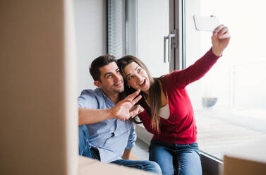 Young couple with cardboard boxes and smartphone moving in a new home, taking selfie. - HPIF30706