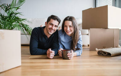 Ein junges, glückliches Paar mit einer Tasse und Kartons, die auf dem Boden liegen, zieht in ein neues Haus ein. - HPIF30721