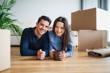 Ein junges, glückliches Paar mit einer Tasse und Kartons, die auf dem Boden liegen, zieht in ein neues Haus ein. - HPIF30722