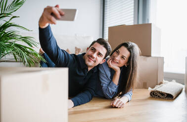 A young happy couple with a smartphone and cardboard boxes lying on a floor, taking selfie when moving in a new home. - HPIF30723