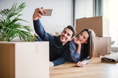 A young happy couple with a smartphone and cardboard boxes lying on a floor, taking selfie when moving in a new home. - HPIF30724