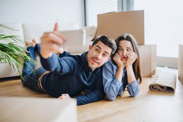 A young happy couple with a smartphone and cardboard boxes lying on a floor, taking selfie and making a face when moving in a new home. - HPIF30725