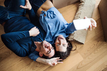 A top view of young happy couple with a smartphone and cardboard boxes lying on a floor, taking selfie when moving in a new home. - HPIF30726