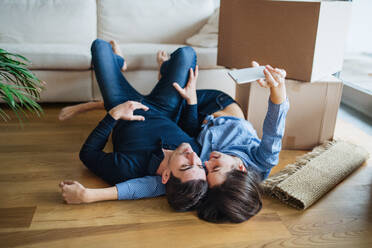 A young happy couple with a smartphone and cardboard boxes lying on a floor, taking selfie when moving in a new home. - HPIF30727