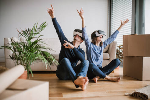Ein junges Paar mit VR-Brillen und Pappkartons sitzt barfuß auf dem Boden und zieht in ein neues Haus ein. - HPIF30728