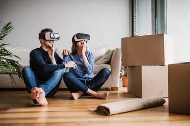 A young couple with VR goggles and cardboard boxes sitting barefoot on a floor, moving in a new home. - HPIF30729