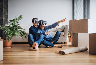 A young couple with VR goggles and cardboard boxes sitting barefoot on a floor, moving in a new home. - HPIF30730