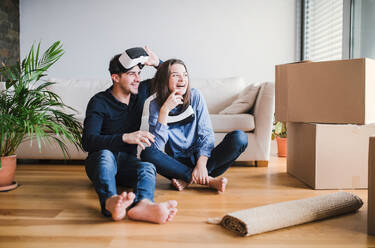 A young couple with VR goggles and cardboard boxes sitting on a floor, moving in a new home. - HPIF30732