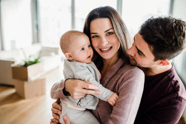 Ein Porträt eines glücklichen jungen Paares mit einem Baby und Kartons beim Umzug in ein neues Haus. - HPIF30734