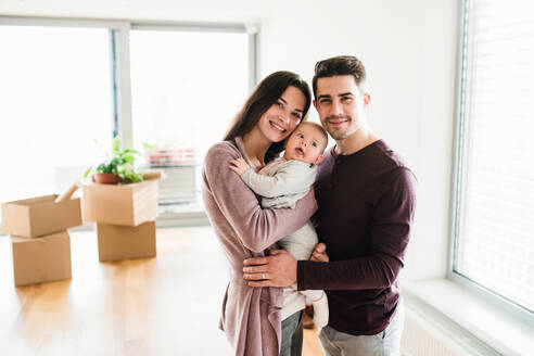 Ein Porträt eines glücklichen jungen Paares mit einem Baby und Kartons beim Umzug in ein neues Haus. - HPIF30735