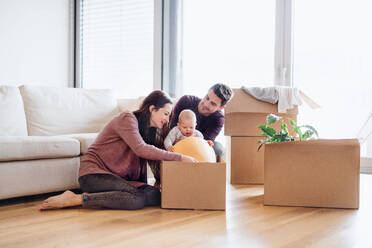 Ein Porträt eines glücklichen jungen Paares, das mit einem Baby und Kartons auf einem Sofa sitzt und in ein neues Haus zieht. - HPIF30755