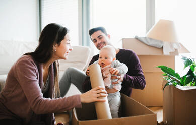 Ein Porträt eines glücklichen jungen Paares, das mit einem Baby und Kartons auf einem Sofa sitzt und in ein neues Haus zieht. - HPIF30757