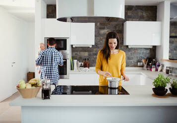 A portrait of young family standing in a kitchen at home, a man holding a baby and a woman cooking. - HPIF30760