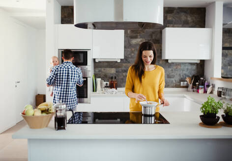 Ein Porträt einer jungen Familie, die zu Hause in einer Küche steht, ein Mann hält ein Baby und eine Frau kocht. - HPIF30760