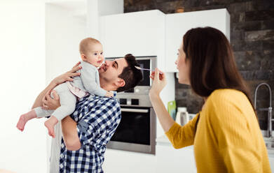 Ein Porträt einer jungen Familie, die zu Hause in einer Küche steht, ein Mann hält ein Baby und eine Frau füttert es mit einem Löffel. - HPIF30761