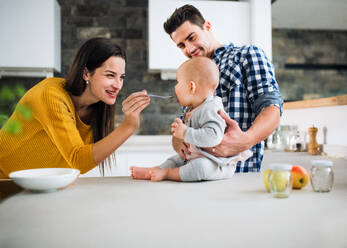 A portrait of young family standing in a kitchen at home, a man holding a baby and a woman feeding her with a spoon. - HPIF30763