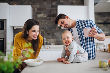 A portrait of young family standing in a kitchen at home, a man holding a baby and a woman feeding her with a spoon. - HPIF30764