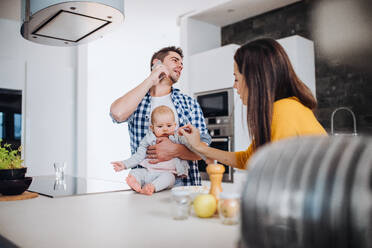 Ein Porträt einer jungen Familie, die zu Hause in einer Küche steht. Ein Mann mit Smartphone hält ein Baby und eine Frau füttert es mit einem Löffel. - HPIF30765