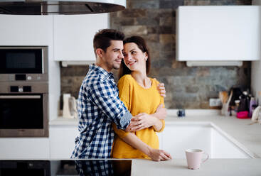 A young happy couple in love standing indoors in a kitchen at home, hugging. - HPIF30769