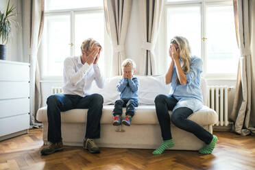 Young parents and toddler son sitting on a sofa inside in a bedroom, covering eyes when playing peekaboo. - HPIF30783