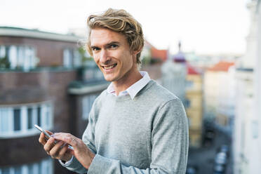 Young man with smartphone standing on a terrace or balcony in a city, texting. - HPIF30787