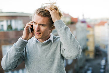 Young man with smartphone standing on a terrace or balcony in a city, making a phone call. - HPIF30789