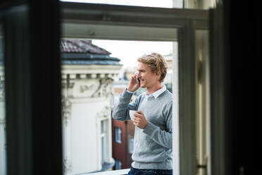 Young man with coffee and smartphone standing on a terrace or balcony in a city, making a phone call. - HPIF30792