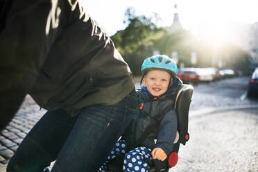 A small toddler boy with helmet sitting in bicycle seat with unrecognizable father outdoors on a street in city. - HPIF30796