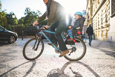 A small toddler boy with helmet sitting in bicycle seat with unrecognizable father outdoors on a street in city. - HPIF30797