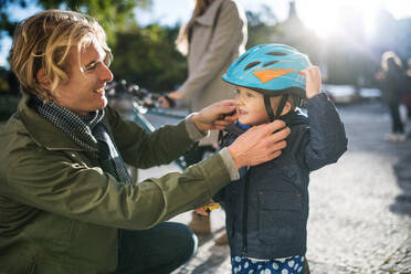 A young father putting on a bike helmet on his toddler son's head outdoors in city. - HPIF30798