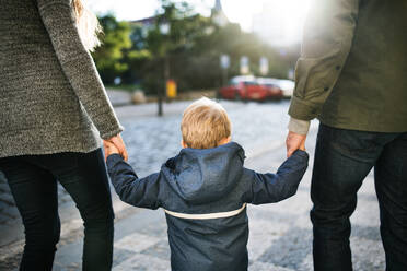 A rear view of small toddler boy with unrecognizable parents walking outdoors in city, holding hands. - HPIF30802