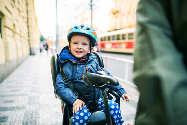 A small toddler boy with helmet sitting in bicycle seat with unrecognizable father outdoors on a street in city. - HPIF30806