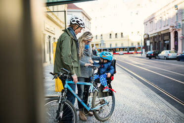 A small toddler boy with helmet sitting in bicycle seat with young parents outdoors on a street in city. - HPIF30807