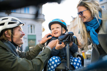 A small toddler boy with bicycle helmet and young parents outdoors in city. - HPIF30808