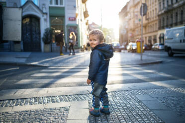 A cute small toddler boy standing by a road outdoors in city at sunset, looking back. - HPIF30809