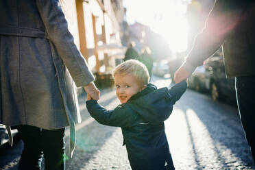 A rear view of small toddler boy with unrecognizable parents walking outdoors in city, holding hands. - HPIF30811