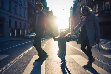 A small toddler boy with parents crossing a road outdoors in city at sunset, holding hands. - HPIF30814