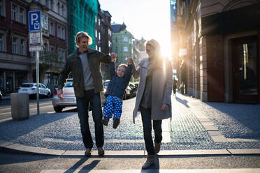 A small toddler boy with parents crossing a road outdoors in city at sunset, holding hands. - HPIF30815