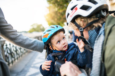 A small toddler boy with bicycle helmet and unrecognizable young parents outdoors in city. - HPIF30826