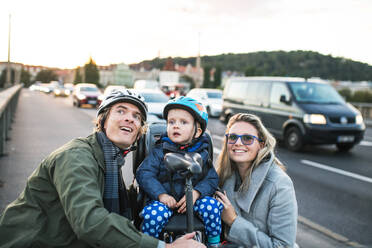 A small toddler boy with helmet sitting in bicycle seat with young parents outdoors on a street in city. - HPIF30827