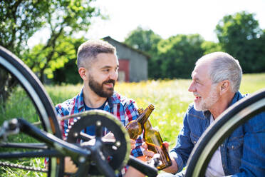 An adult hipster son and senior father repairing bicycle outside on a sunny summer day, drinking beer. - HPIF30909