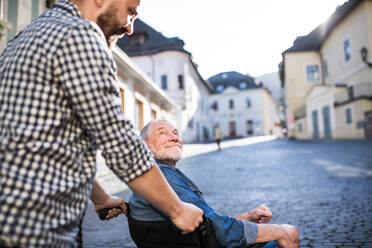 An unrecognizable adult hipster son with his senior father in wheelchair on a walk in town. - HPIF30925