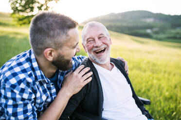 An adult hipster son with his senior father in wheelchair on a walk on a meadow in nature at sunset, laughing. Close up. - HPIF30933