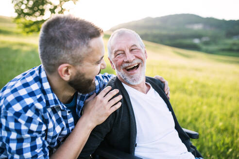 An adult hipster son with his senior father in wheelchair on a walk on a meadow in nature at sunset, laughing. Close up. - HPIF30933