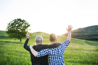 An adult hipster son with his senior father on a walk in nature at sunset, arms around each other. Rear view. - HPIF30944