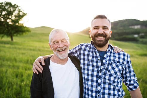A portrait of a laughing adult hipster son with senior father in nature at sunset, arms around each other. - HPIF30948