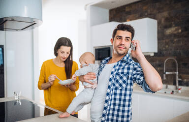 A portrait of young family standing in a kitchen at home, a man making a phone call and a woman feeding a baby. - HPIF31037